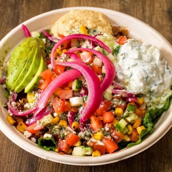 Colorful salad bowl with avocado, pickled onions, hummus, tzatziki, and chopped vegetables on wood.