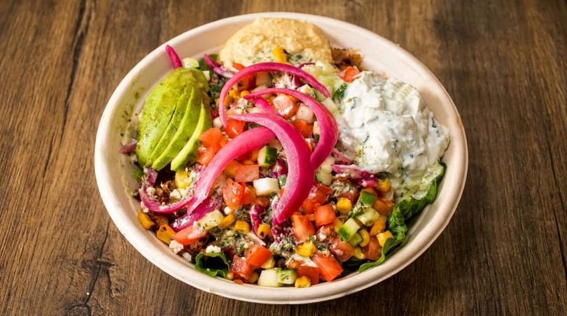 Colorful salad bowl with avocado, pickled onions, hummus, tzatziki, and chopped vegetables on wood.